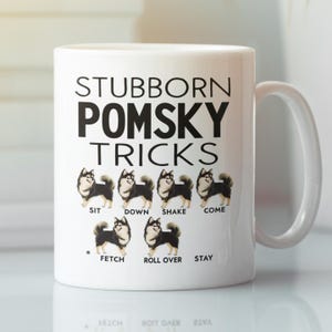 Op de afbeelding: Een witte keramische koffiemok met een zwart-witte illustratie van een Pomsky-hondenras. De mok heeft de tekst "Stubborn Pomsky Tricks" en somt verschillende hondentrucs op met illustraties van de hond die elke truc uitvoert.