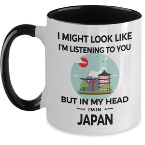 May include: White ceramic coffee mug with a black handle and black rim. The mug has a graphic of a Japanese cityscape with the text "I might look like I'm listening to you, but in my head I'm in Japan."