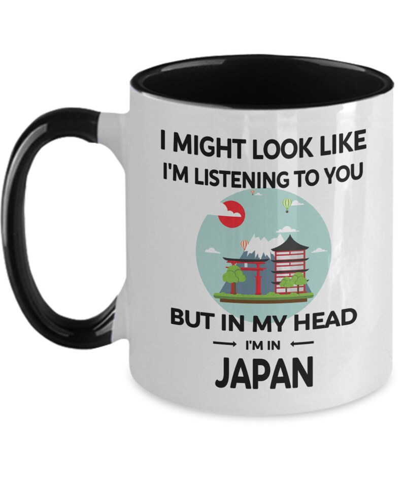 May include: White ceramic coffee mug with a black handle and black rim. The mug has a graphic of a Japanese cityscape with the text "I might look like I'm listening to you, but in my head I'm in Japan."