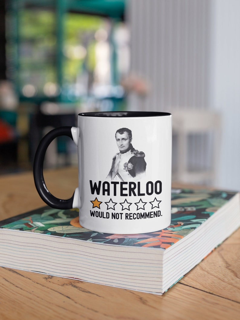 May include: White ceramic mug with a black handle and black rim. The mug features a black and white image of Napoleon Bonaparte and the text "Waterloo" with a one-star rating and the text "Would not recommend."