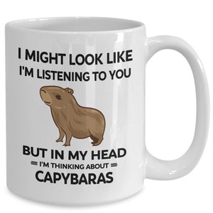 K&ouml;nnte beinhalten: Eine wei&szlig;e Keramik-Kaffeetasse mit einer schwarzen Grafik eines Capybaras und dem Text "I might look like I'm listening to you but in my head = I'm thinking about = capybaras"