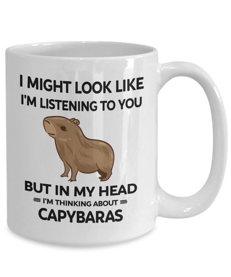 K&ouml;nnte beinhalten: Eine wei&szlig;e Keramik-Kaffeetasse mit einer schwarzen Grafik eines Capybaras und dem Text "I might look like I'm listening to you but in my head = I'm thinking about = capybaras"