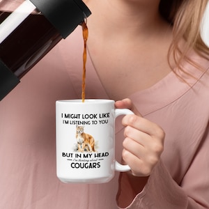 May include: White ceramic coffee mug with a black graphic of a cougar and the text "I might look like I'm listening to you, but in my head I'm thinking about cougars."