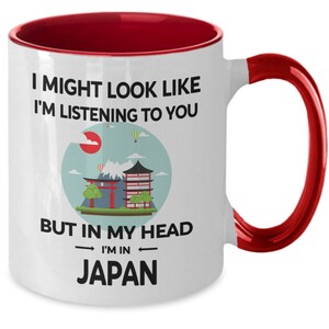 May include: White ceramic coffee mug with a red rim and handle. The mug features a graphic of a Japanese cityscape with the text "I might look like I'm listening to you, but in my head I'm in Japan."
