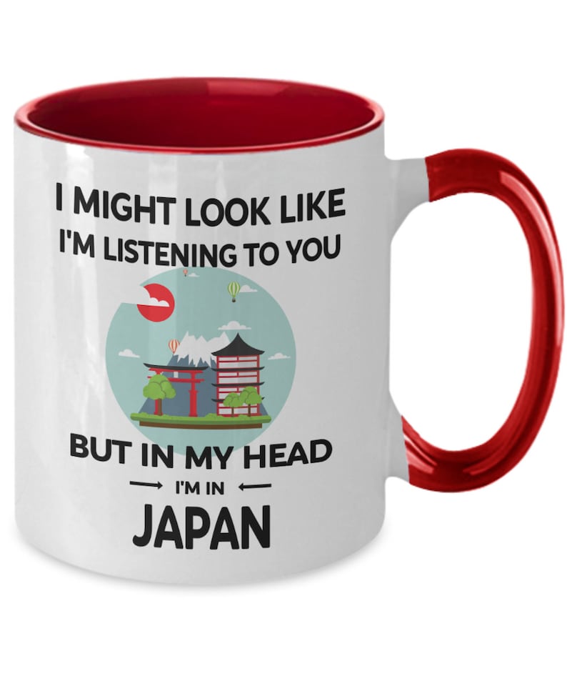 May include: White ceramic coffee mug with a red rim and handle. The mug features a graphic of a Japanese cityscape with the text "I might look like I'm listening to you, but in my head I'm in Japan."