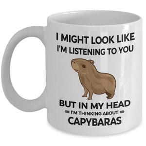K&ouml;nnte beinhalten: Eine wei&szlig;e Keramik-Kaffeetasse mit einer schwarzen und braunen Illustration eines Capybaras. Der Text auf der Tasse lautet "I might look like I'm listening to you, but in my head = I'm thinking about = Capybaras."