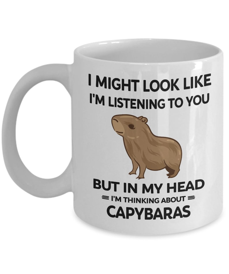 K&ouml;nnte beinhalten: Eine wei&szlig;e Keramik-Kaffeetasse mit einer schwarzen und braunen Illustration eines Capybaras. Der Text auf der Tasse lautet "I might look like I'm listening to you, but in my head = I'm thinking about = Capybaras."
