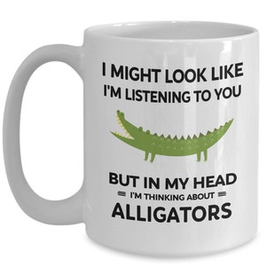 K&ouml;nnte beinhalten: Eine wei&szlig;e Keramik-Kaffeetasse mit einem schwarzen und gr&uuml;nen Alligator-Motiv. Die Tasse tr&auml;gt den Text "Ich sehe vielleicht aus, als w&uuml;rde ich zuh&ouml;ren, aber in meinem Kopf = denke ich an = Alligatoren."