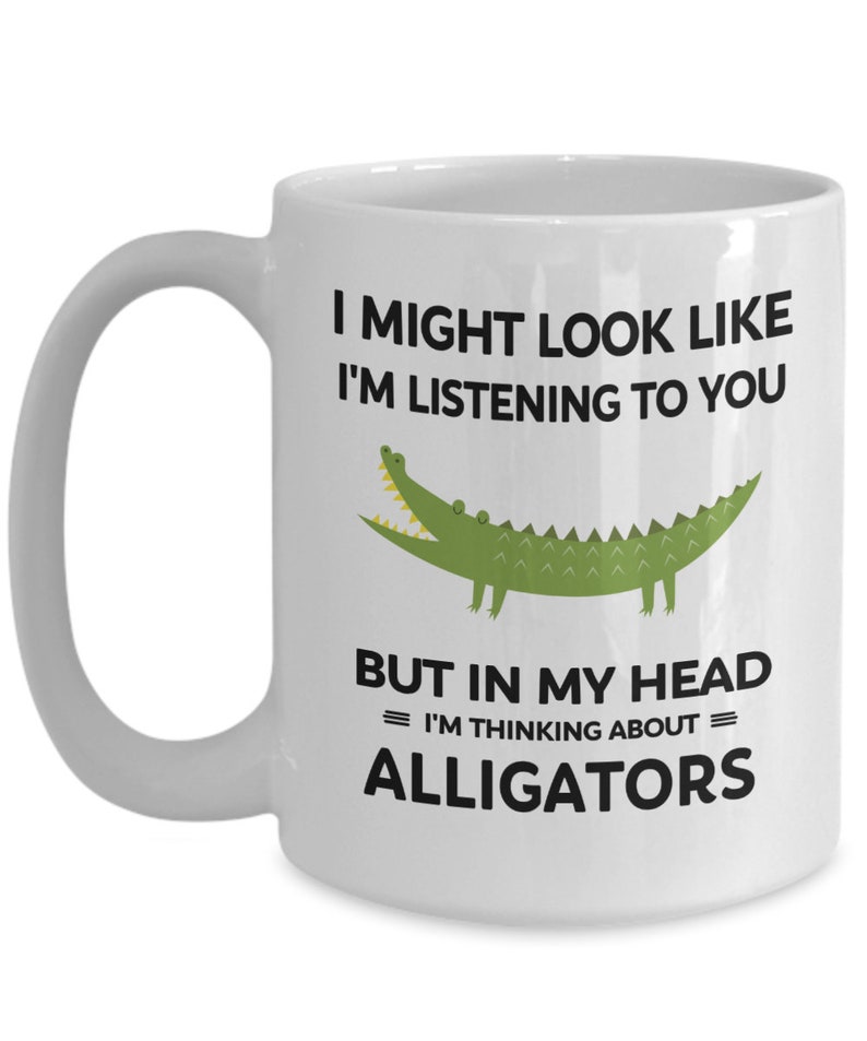 K&ouml;nnte beinhalten: Eine wei&szlig;e Keramik-Kaffeetasse mit einem schwarzen und gr&uuml;nen Alligator-Motiv. Die Tasse tr&auml;gt den Text "Ich sehe vielleicht aus, als w&uuml;rde ich zuh&ouml;ren, aber in meinem Kopf = denke ich an = Alligatoren."
