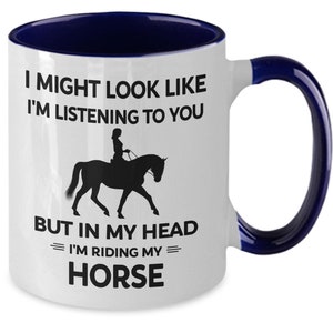 May include: White ceramic coffee mug with a blue rim and handle. The mug features a black silhouette of a person riding a horse with the text "I might look like I'm listening to you but in my head I'm riding my horse."