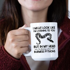 Op de afbeelding: Witte keramische mok met de tekst "I MIGHT LOOK LIKE I'M LISTENING TO YOU BUT IN MY HEAD I'm thinking about Burmese Pythons" en een zwart-witte slangenillustratie.