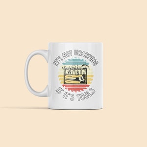 May include: White ceramic mug with a vintage-style graphic that reads "It's not hoarding if it's tools". The graphic features a toolbox with various tools inside, surrounded by a circular saw blade design.