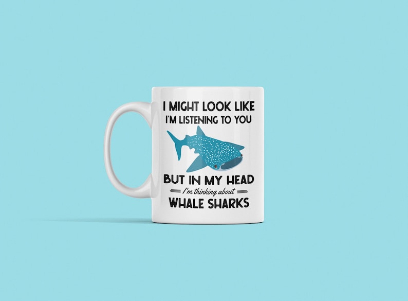 Pu&ograve; includere: Una tazza da caff&egrave; in ceramica bianca con una grafica nera e blu di uno squalo balena. Il testo sulla tazza recita "Potrei sembrare che ti stia ascoltando, ma nella mia testa sto pensando agli squali balena."