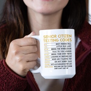 May include: White ceramic mug with the text "SENIOR CITIZEN TEXTING CODES" in gold lettering. The mug lists common texting acronyms and their meanings, such as "LOL" and "BTW". The mug is held by a person.