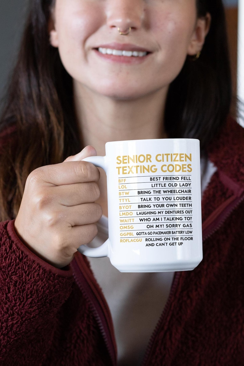 May include: White ceramic mug with the text "SENIOR CITIZEN TEXTING CODES" in gold lettering. The mug lists common texting acronyms and their meanings, such as "LOL" and "BTW". The mug is held by a person.