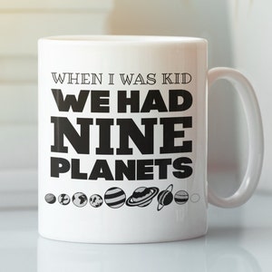 Puede incluir: Taza de cerámica blanca con texto negro que dice "Cuando era niño, teníamos nueve planetas". La taza tiene un contorno negro del sistema solar con nueve planetas.