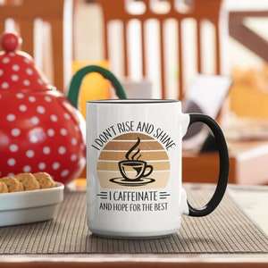 Op de afbeelding: Witte keramische koffiemok met een zwarte handgreep. De mok heeft een afbeelding van een zonsondergang met een kopje koffie en de tekst "I don't rise and shine = I caffeinate = and hope for the best".