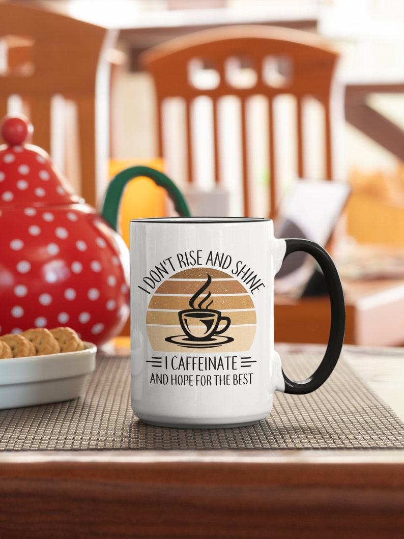 Op de afbeelding: Witte keramische koffiemok met een zwarte handgreep. De mok heeft een afbeelding van een zonsondergang met een kopje koffie en de tekst "I don't rise and shine = I caffeinate = and hope for the best".