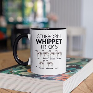 Op de afbeelding: Witte keramische koffiemok met een zwarte handgreep en zwarte rand. De mok heeft een zwarte afbeelding met de tekst "Stubborn Whippet Tricks" met een lijst van hondentrucs en een silhouet van een Whippet-hond voor elke truc.