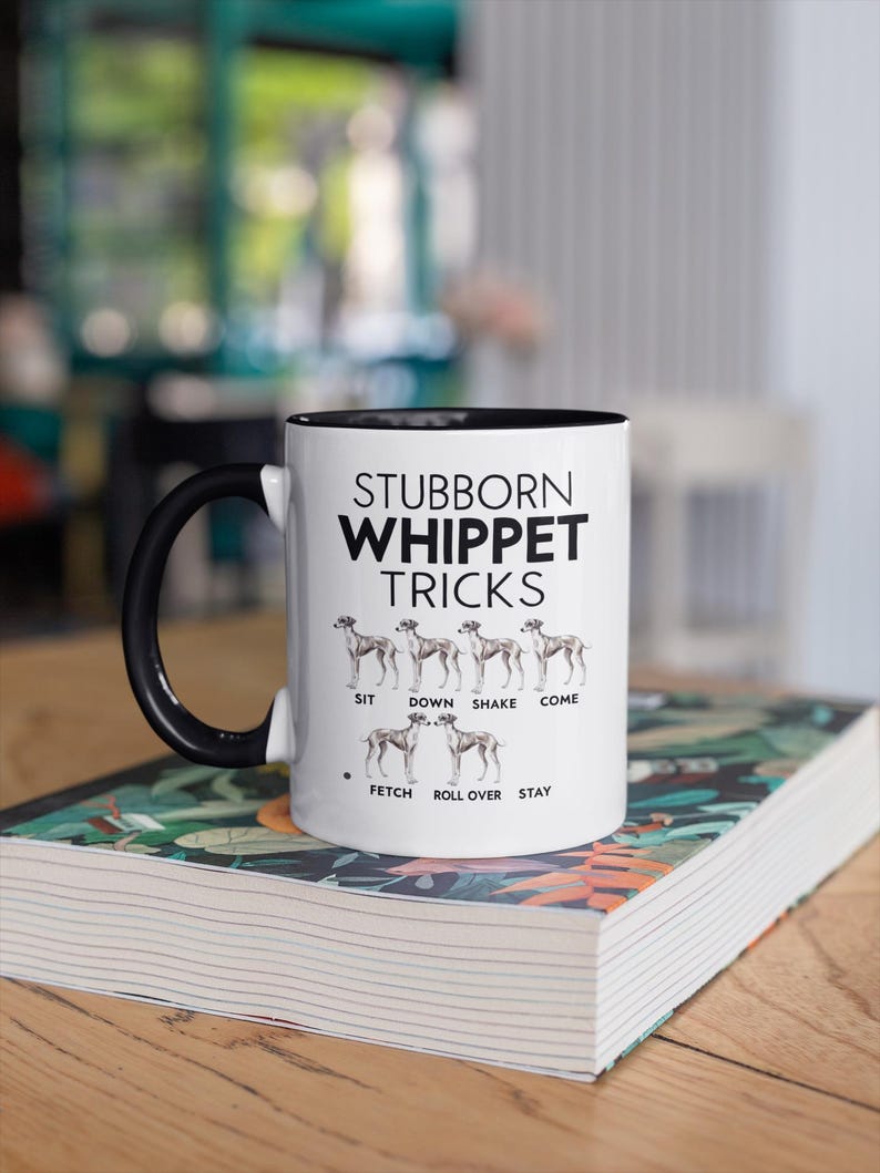 Op de afbeelding: Witte keramische koffiemok met een zwarte handgreep en zwarte rand. De mok heeft een zwarte afbeelding met de tekst "Stubborn Whippet Tricks" met een lijst van hondentrucs en een silhouet van een Whippet-hond voor elke truc.