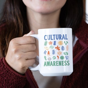 May include: White ceramic mug with a blue and green design featuring the words "Cultural Awareness" and a variety of colourful, stylised germs.