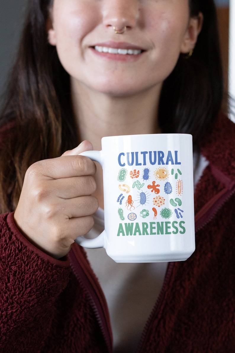 May include: White ceramic mug with a blue and green design featuring the words "Cultural Awareness" and a variety of colourful, stylised germs.