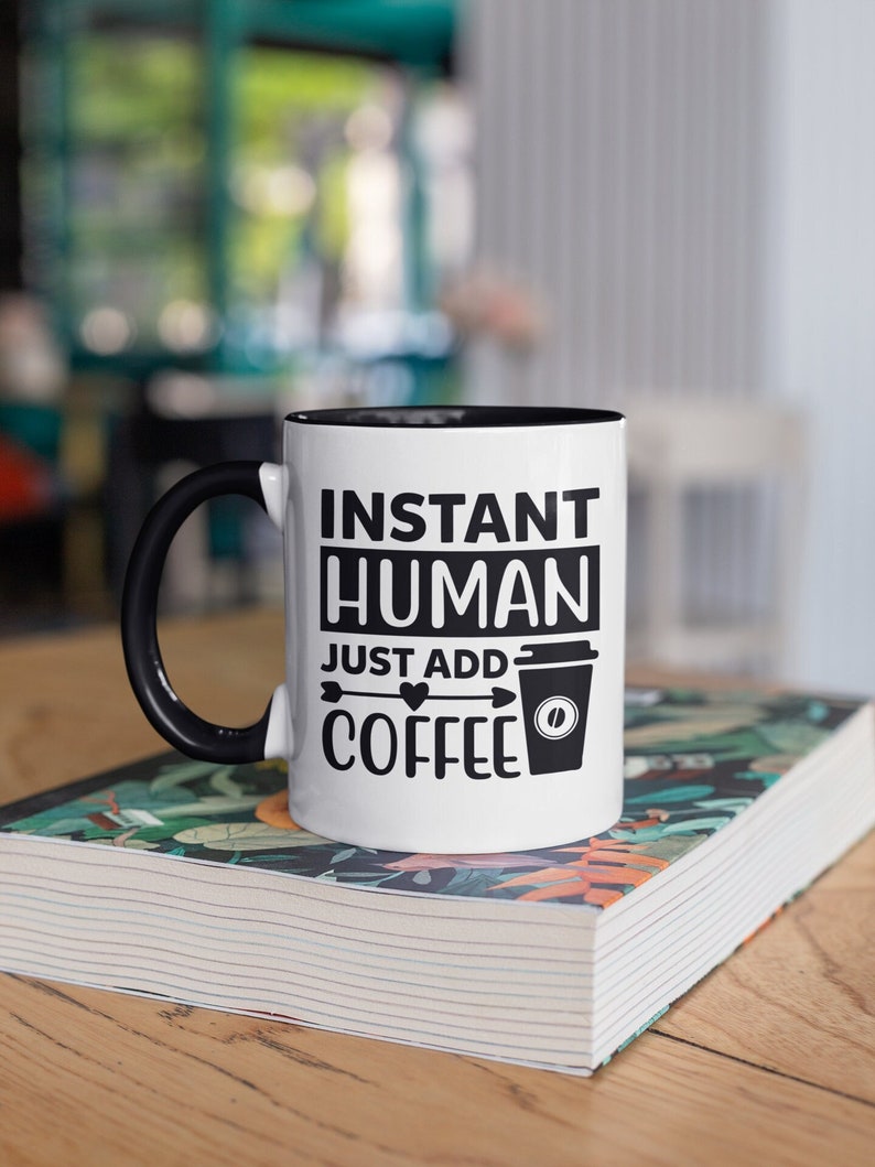 May include: White coffee mug with a black handle and black rim. The mug has black text that reads "Instant Human Just Add Coffee" with a coffee cup icon and a heart.
