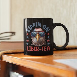 May include: Black coffee mug with a bald eagle wearing sunglasses and a red, white, and blue bandana. The text on the mug reads "Sippin' on Liber-Tea".