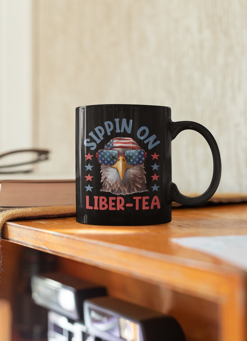May include: Black coffee mug with a bald eagle wearing sunglasses and a red, white, and blue bandana. The text on the mug reads "Sippin' on Liber-Tea".