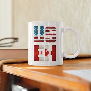 May include: White ceramic mug with the letters "US" and "CA" printed in red, white, and blue. The letters are stylized to look like the American and Canadian flags.