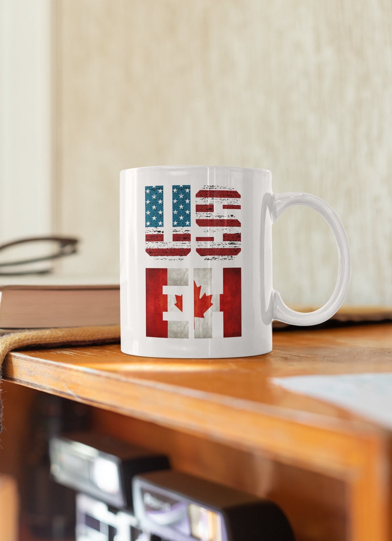 May include: White ceramic mug with the letters "US" and "CA" printed in red, white, and blue. The letters are stylized to look like the American and Canadian flags.