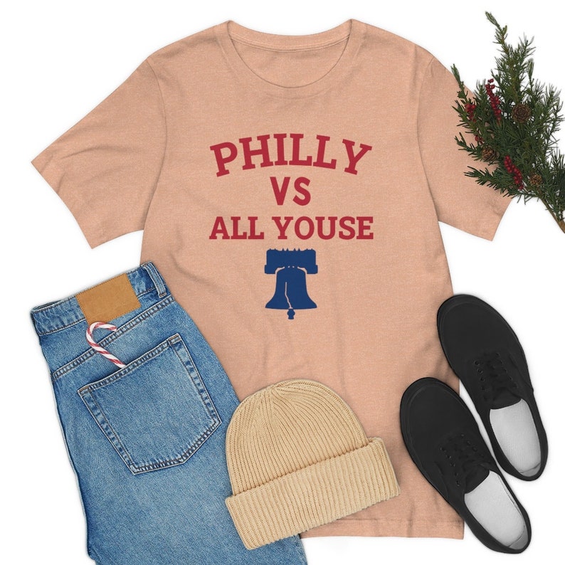 May include: A peach-colored t-shirt with the text "PHILLY VS ALL YOUSE" in red and blue lettering. A blue and white Liberty Bell graphic is below the text.