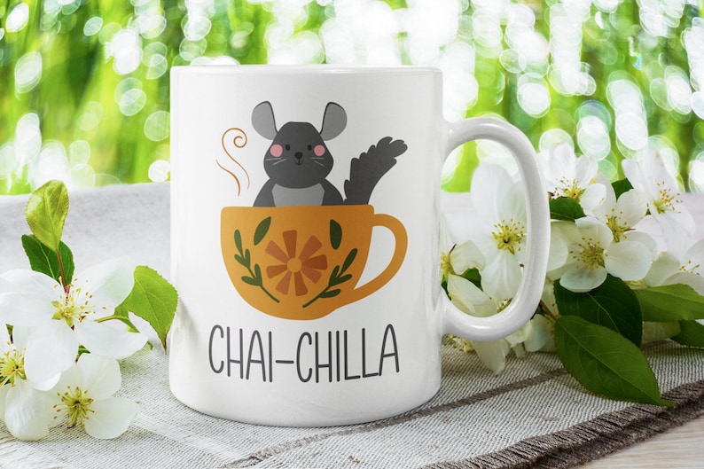 May include: White ceramic mug featuring a black and grey cartoon chinchilla sitting in a yellow teacup with a floral design. The text "CHAI-CHILLA" is printed on the mug.