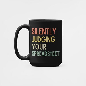 K&ouml;nnte beinhalten: Schwarze Kaffeetasse mit dem Text "SILENTLY JUDGING YOUR SPREADSHEET" in einem Retro-Farbschema.