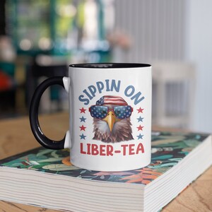 May include: A white ceramic mug with a black rim and handle. The mug features a cartoon bald eagle wearing sunglasses and a red, white, and blue hat. The text "Sippin' on Liber-Tea" is printed on the mug.