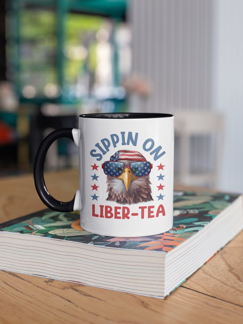May include: A white ceramic mug with a black rim and handle. The mug features a cartoon bald eagle wearing sunglasses and a red, white, and blue hat. The text "Sippin' on Liber-Tea" is printed on the mug.