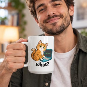 Op de afbeelding: Een witte keramische mok met een cartoonillustratie van een oranje cyperse kat die achter een blauwe laptop zit. De kat heeft grote groene ogen en kijkt omhoog. Onder de kat staat het woord "what?" in dikgedrukte, zwarte letters.