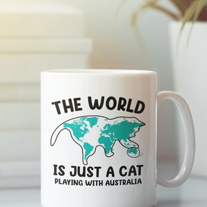 May include: White ceramic mug with a black handle. The mug features the text "THE WORLD IS JUST A CAT PLAYING WITH AUSTRALIA" with a teal map of the world shaped like a cat.