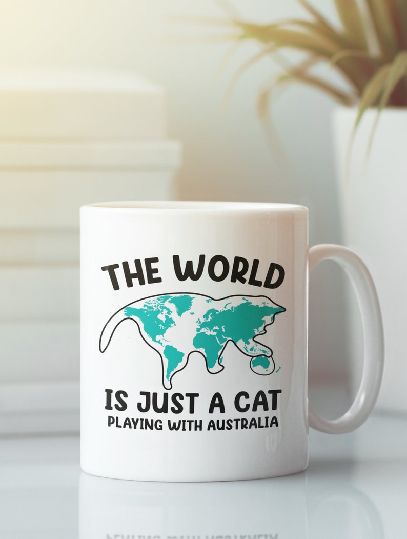 May include: White ceramic mug with a black handle. The mug features the text "THE WORLD IS JUST A CAT PLAYING WITH AUSTRALIA" with a teal map of the world shaped like a cat.