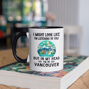 May include: White coffee mug with a black handle and black rim. The mug has a graphic of a city skyline with mountains and a sun. The text on the mug reads "I might look like I'm listening to you but in my head I'm in Vancouver."
