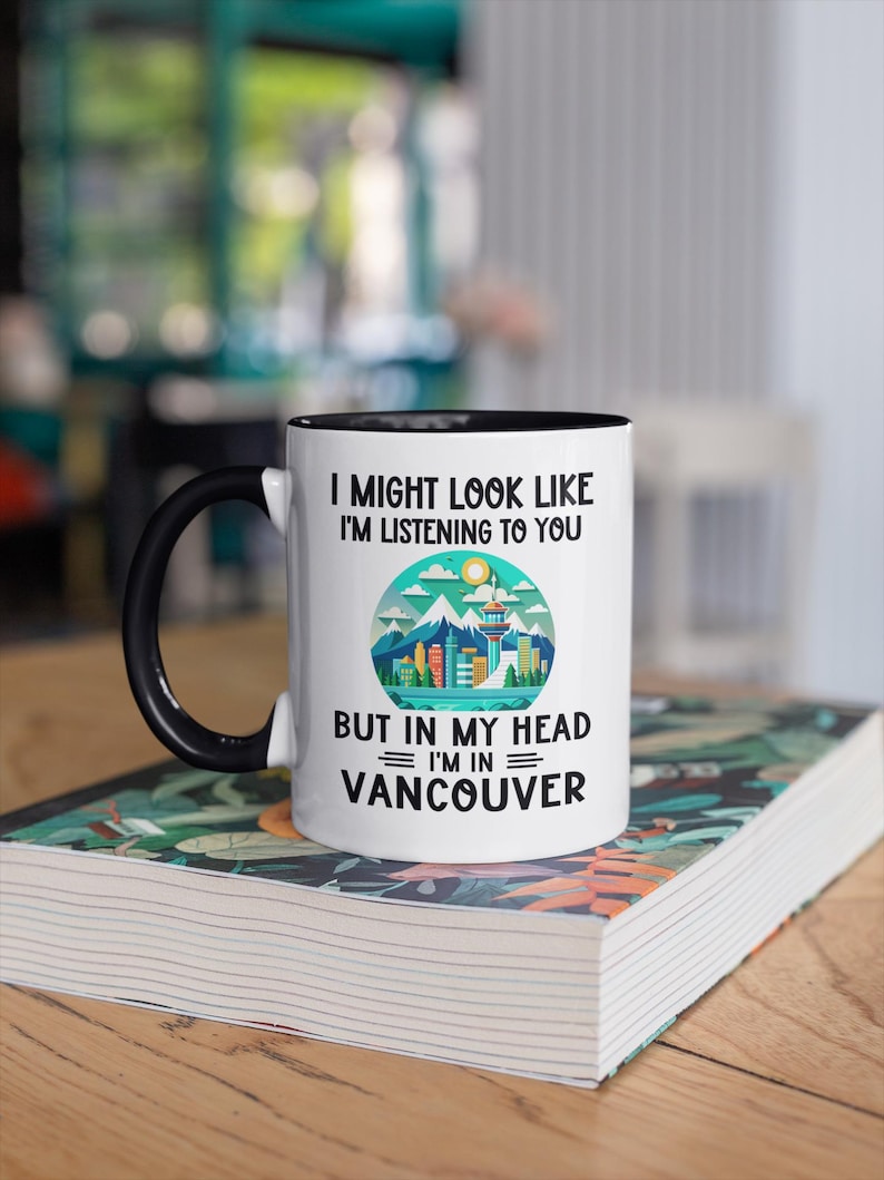 May include: White coffee mug with a black handle and black rim. The mug has a graphic of a city skyline with mountains and a sun. The text on the mug reads "I might look like I'm listening to you but in my head I'm in Vancouver."