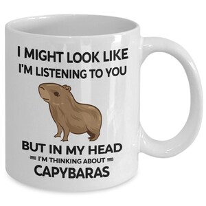 K&ouml;nnte beinhalten: Eine wei&szlig;e Keramik-Kaffeetasse mit einer schwarzen und braunen Illustration eines Capybaras. Die Tasse tr&auml;gt den Text "I might look like I'm listening to you, but in my head = I'm thinking about = Capybaras".