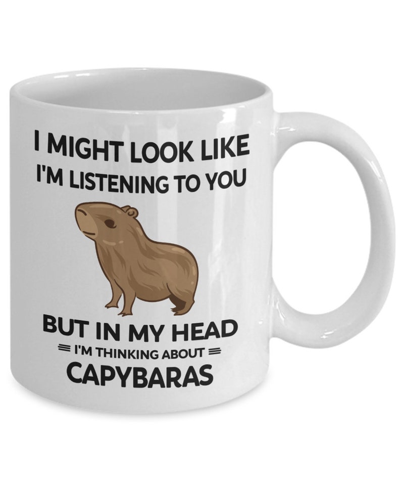 K&ouml;nnte beinhalten: Eine wei&szlig;e Keramik-Kaffeetasse mit einer schwarzen und braunen Illustration eines Capybaras. Die Tasse tr&auml;gt den Text "I might look like I'm listening to you, but in my head = I'm thinking about = Capybaras".