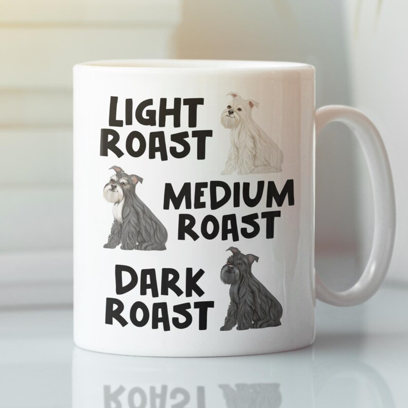 May include: White ceramic coffee mug with a black graphic of three schnauzers of different colors. The text "Light Roast", "Medium Roast", and "Dark Roast" is printed on the mug.