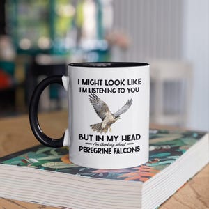 May include: White coffee mug with a black rim and handle. The mug features a black and white graphic of a peregrine falcon with the text "I might look like I'm listening to you, but in my head I'm thinking about Peregrine Falcons."