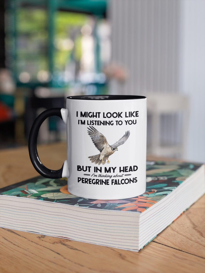 May include: White coffee mug with a black rim and handle. The mug features a black and white graphic of a peregrine falcon with the text "I might look like I'm listening to you, but in my head I'm thinking about Peregrine Falcons."