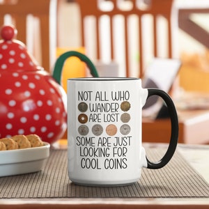 May include: White ceramic mug with a black handle. The mug has a black rim and a black line around the bottom. The mug has a quote that reads "Not all who wander are lost. Some are just looking for cool coins." with a row of coins above the first line of text and a row of coins below the second line of text.
