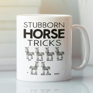 以下が含まれることがあります： 「Stubborn Horse Tricks」と黒文字で書かれた白いセラミック製のコーヒーカップ。イラストには、6頭の漫画の馬が描かれており、それぞれの下に「Sit」「Down」「Shake」「Come」「Fetch」「Roll Over」「Stay」というコマンドが書かれている。