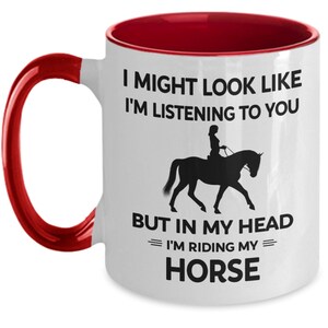 May include: White ceramic coffee mug with a red rim and handle. The mug features a black silhouette of a person riding a horse with the text "I might look like I'm listening to you but in my head I'm riding my horse."