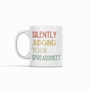 K&ouml;nnte beinhalten: Wei&szlig;e Keramik-Kaffeetasse mit dem Text "SILENTLY JUDGING YOUR SPREADSHEET" in einem Retro-Farbschema.